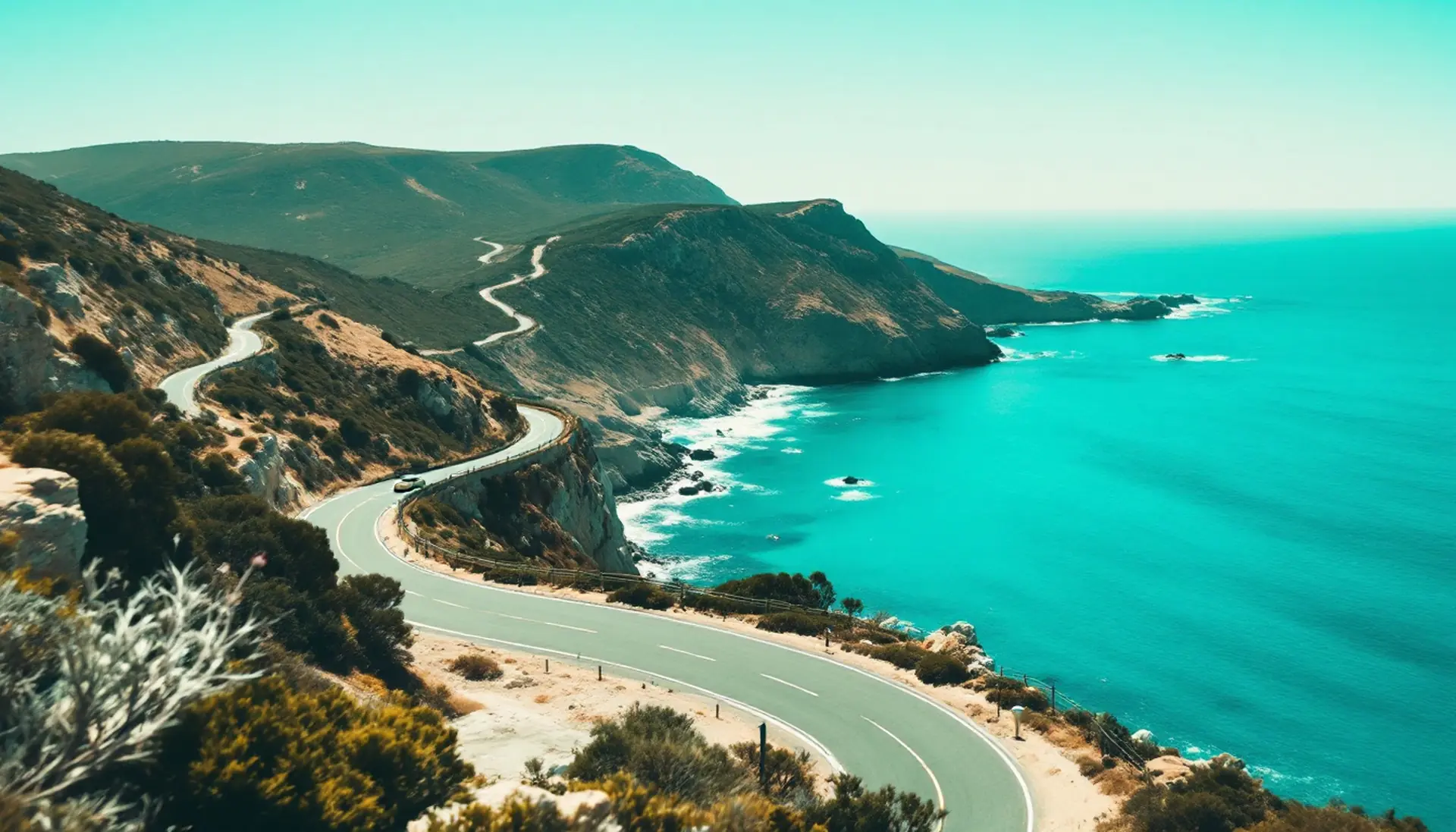 A small cream sedan on a winding coastal road above a turquoise sea